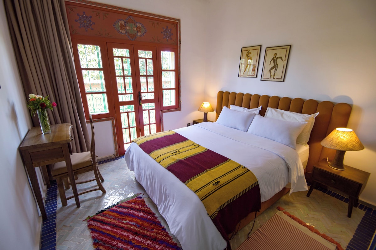Une chambre à coucher comprenant un lit, des tables de chevet, un bureau, une chaise, une fenêtre ornée de ferronnerie décorative et un tapis.
