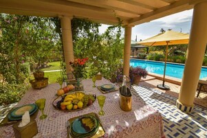 An outdoor dining area with a pool view, featuring a table set with fruits, glasses, and a bottle.