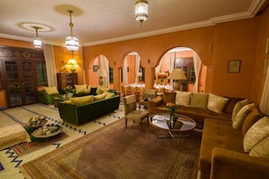 A spacious living room with a green sofa, beige armchairs, and a coffee table. There are two chandeliers, a patterned rug, and a view through an arched doorway.