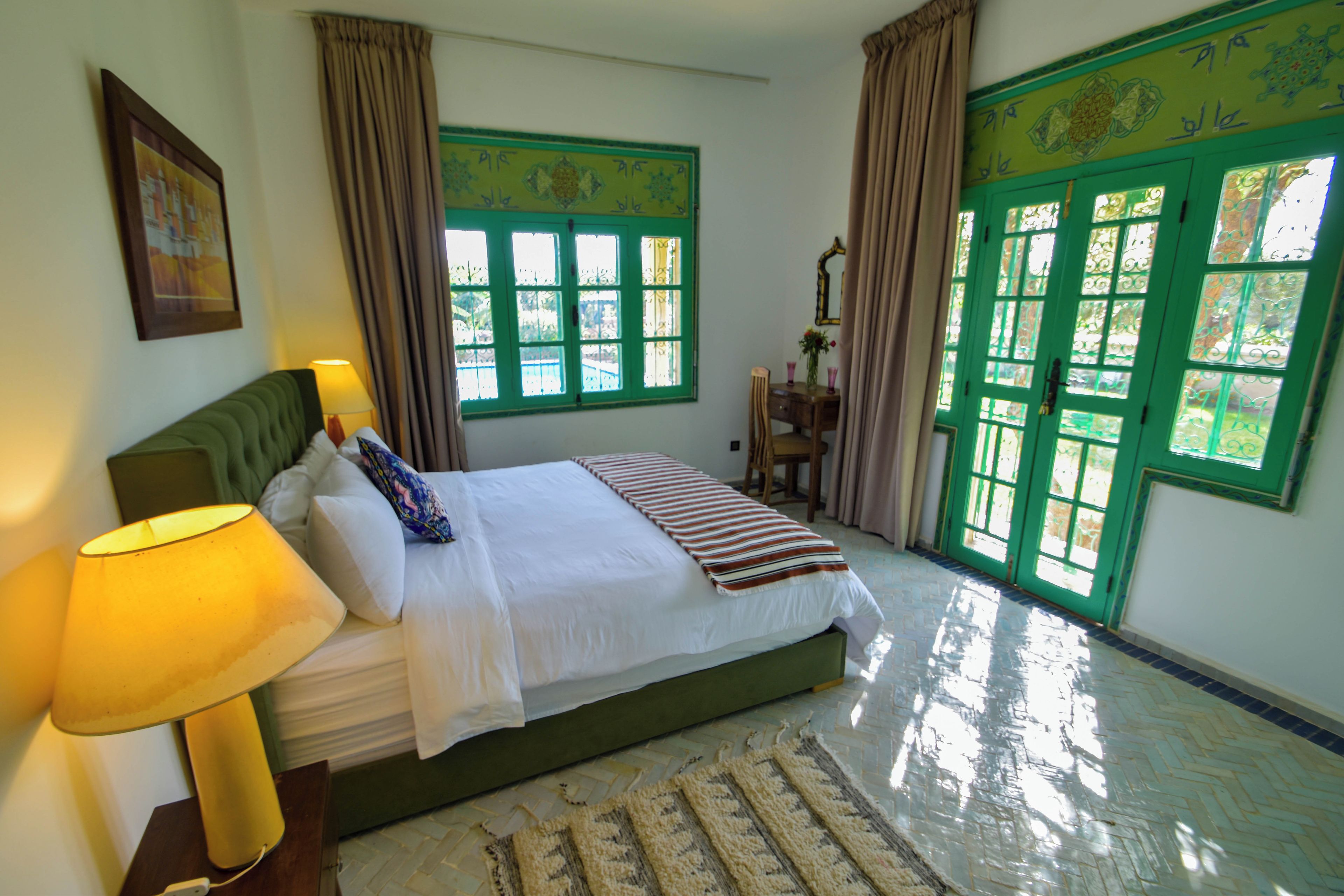 Un dormitorio con cama, mesita de noche, lámpara, cómoda y ventana con contraventanas verdes.