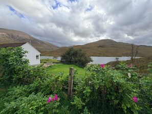A rural landscape with a white building, a fenced area with green vegetation, and a body of water in the background. - The Cottage at Lundavra Farm (Lundavra)