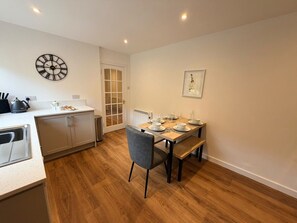 A modern kitchen with a dining area, wooden flooring, and a clock on the wall. - The Cottage at Lundavra Farm (Lundavra)