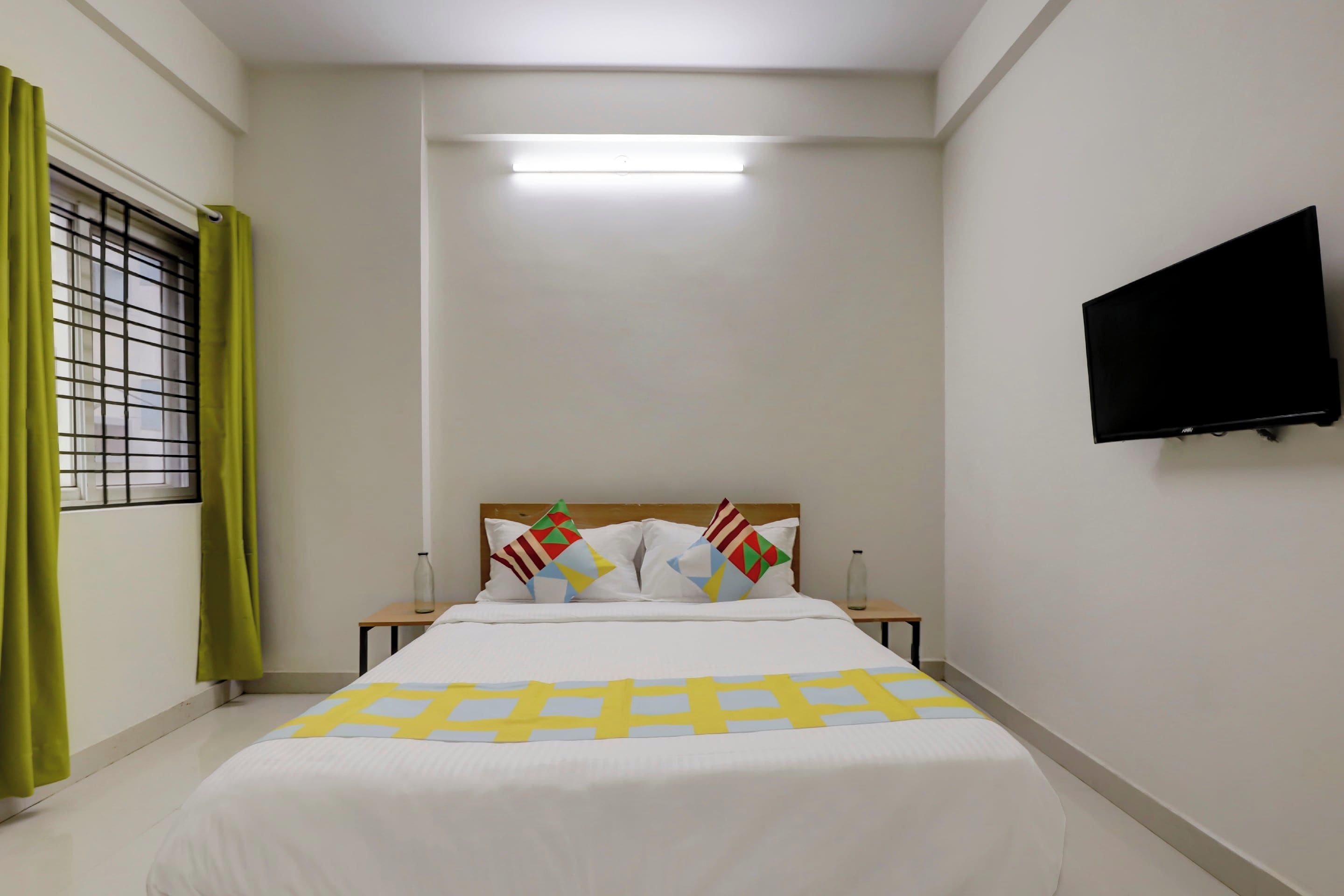 A hotel room with a bed, a flat-screen TV mounted on the wall, a window with green curtains, and a bedside table with a bottle.