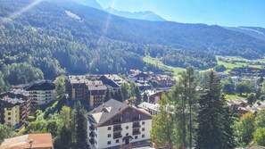 Une ville de montagne avec des bâtiments, dont un hôtel à plusieurs étages et doté de balcons, entourée de forêts denses et surplombant un ciel dégagé.