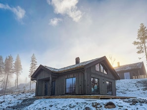 A wooden cabin with a gabled roof, large windows, and a chimney, surrounded by snow and trees.