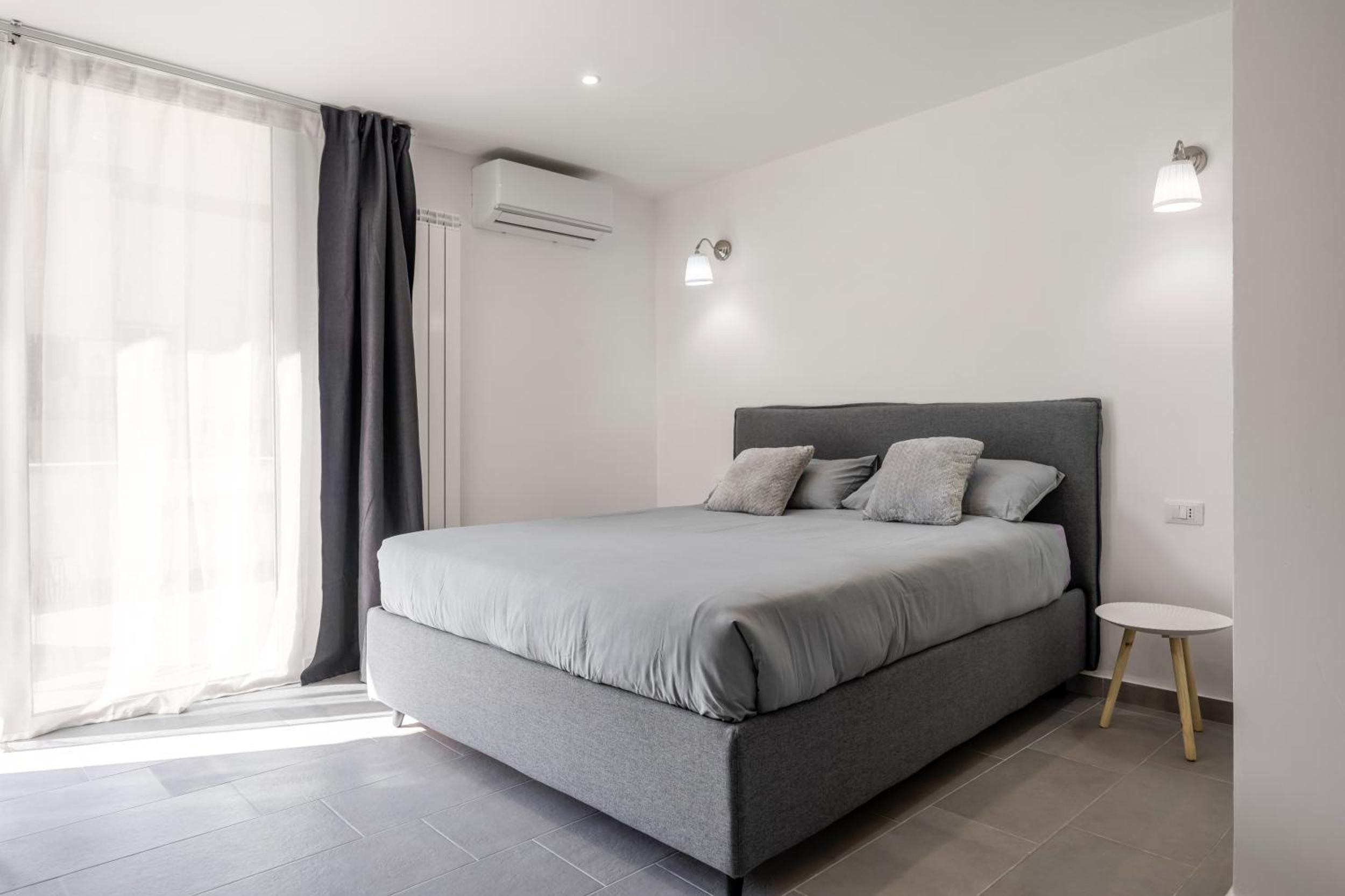 Une chambre moderne avec un lit gris, une petite table ronde et une fenêtre avec des rideaux.