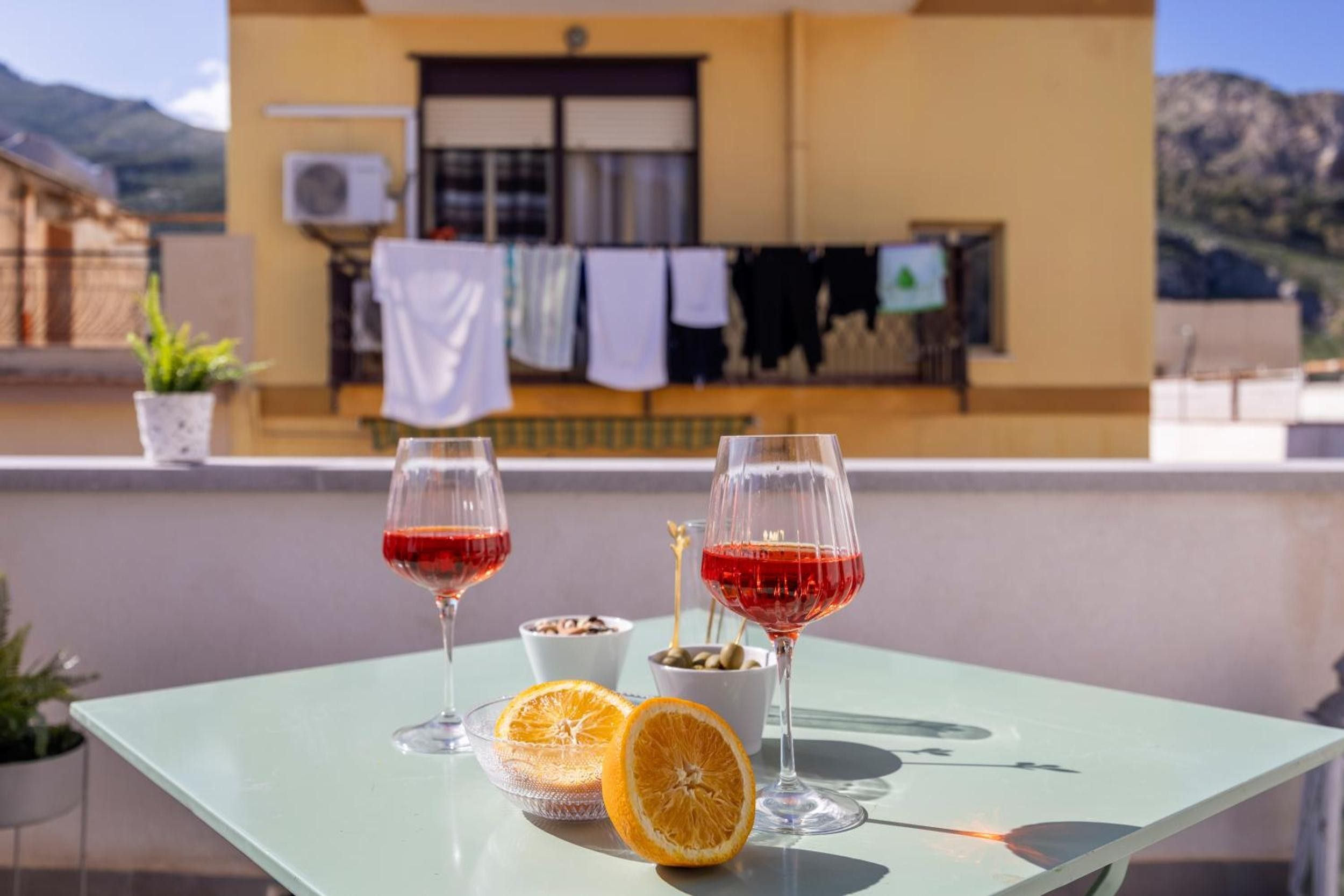 Une table avec deux verres à vin, un bol d’olives et une orange coupée en deux, sur un balcon d’où l’on aperçoit un immeuble et des montagnes.