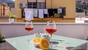 Une table avec deux verres à vin, un bol d’olives et une orange coupée en deux, sur un balcon d’où l’on aperçoit un immeuble et des montagnes.