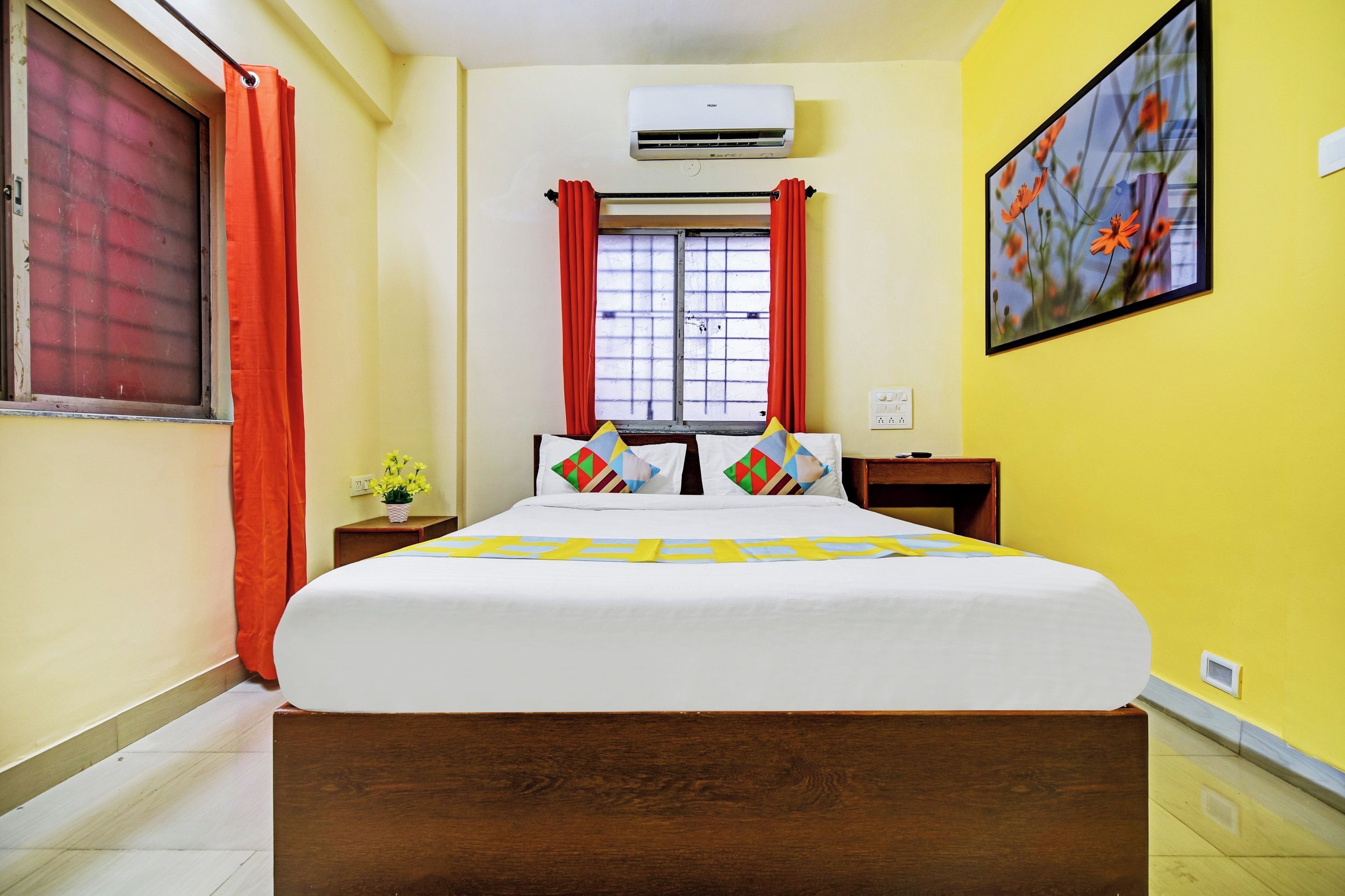 A hotel room with a bed, an air conditioner, a window with curtains, a bedside table with a lamp, and a wall-mounted painting.