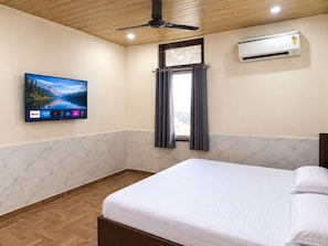 A hotel room with a bed, a ceiling fan, an air conditioning unit, a TV mounted on the wall, and a window with curtains. - Hotel Shubh Laxmi (Jaipur)