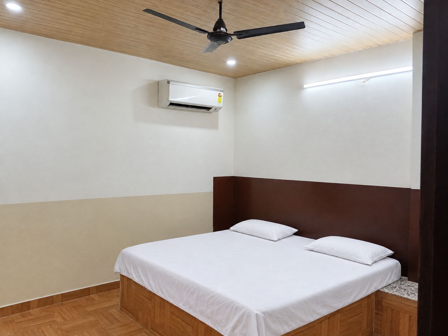 A bedroom with a bed, a ceiling fan, an air conditioning unit, and a wooden floor.