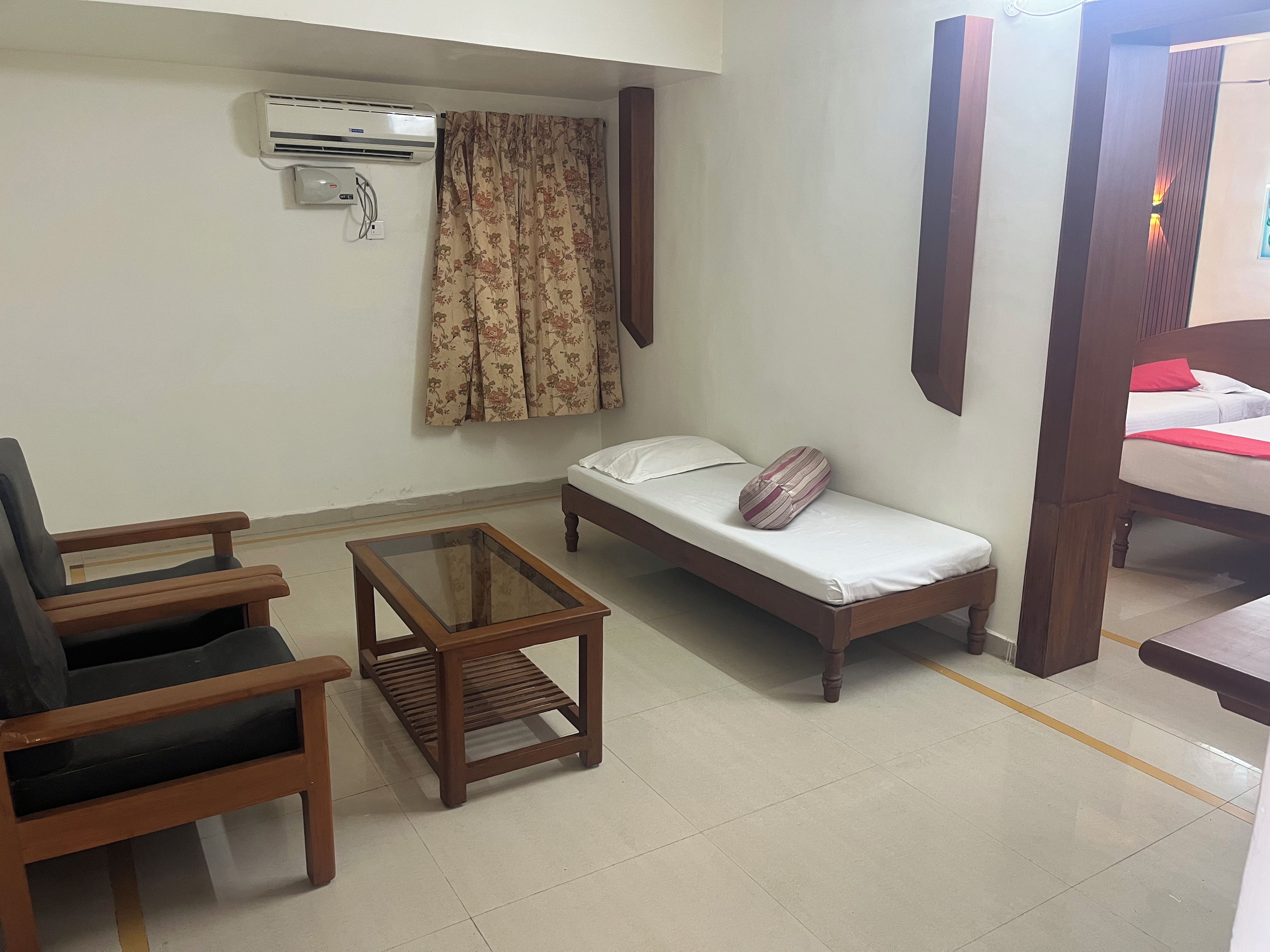 A small, furnished room with a bed, a chair, a small table, and an air conditioning unit.