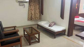 A small, furnished room with a bed, a chair, a small table, and an air conditioning unit.