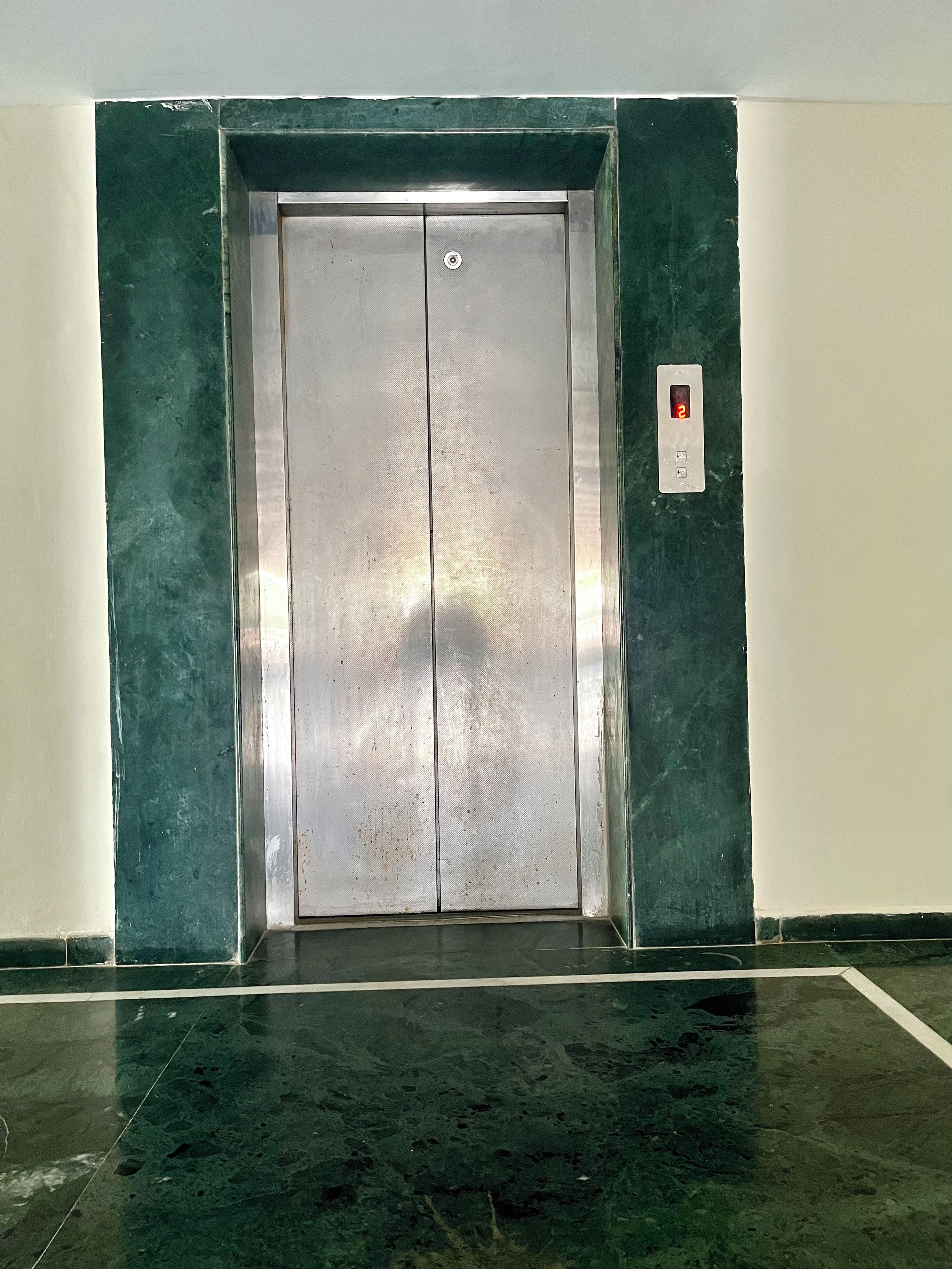 Un ascensor amb acabat metàl·lic, un panell de control amb pantalla digital i una paret amb rajoles verdes.