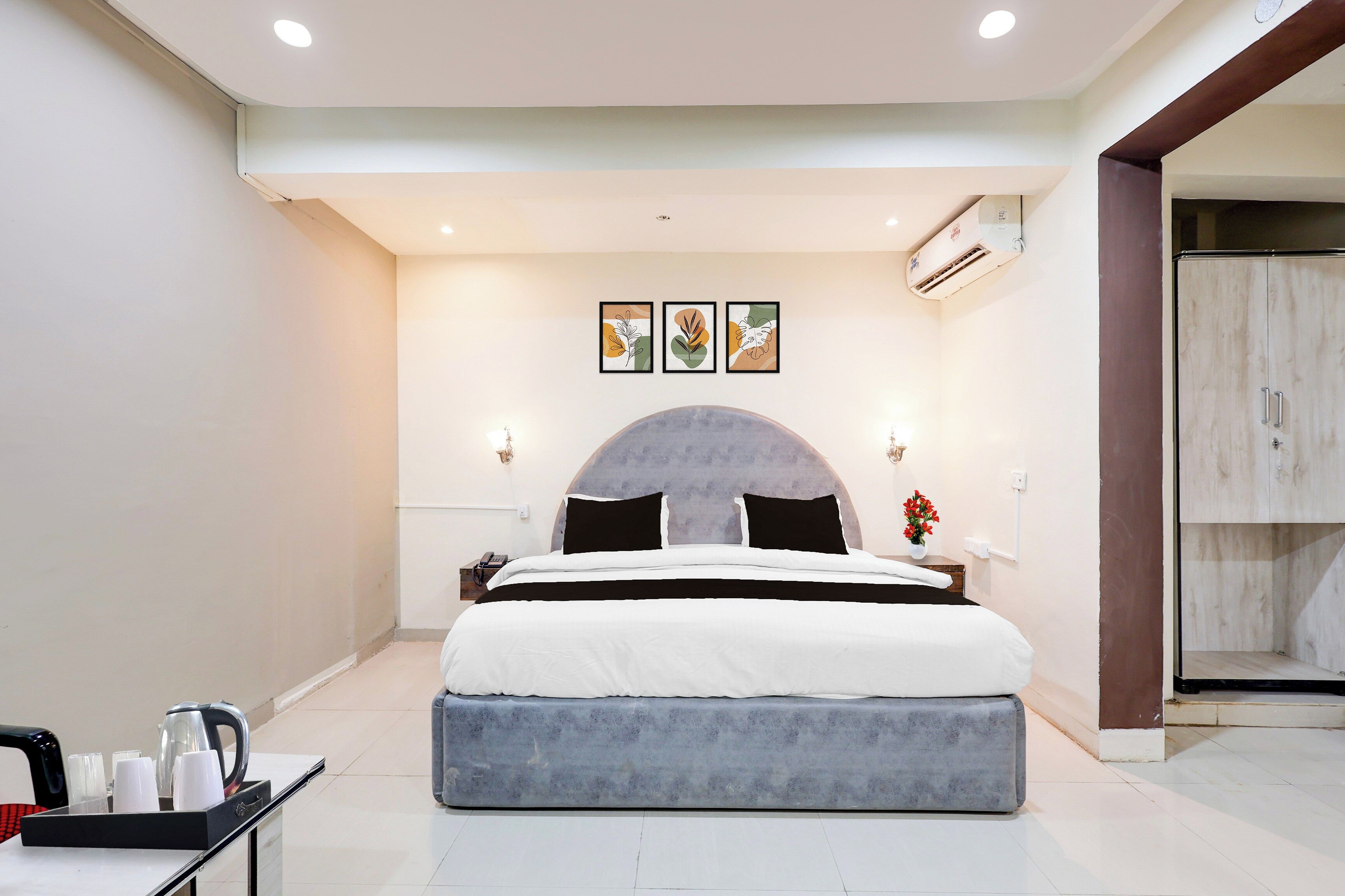 A modern hotel room with a large bed, a bedside table with a kettle, and a small table with cups.