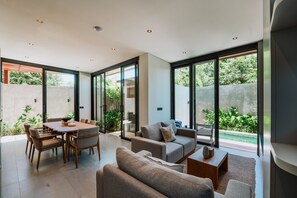 A modern living room with a dining area, large sliding glass doors, and a view of greenery outside. - Bellana Villas (Jimbaran)