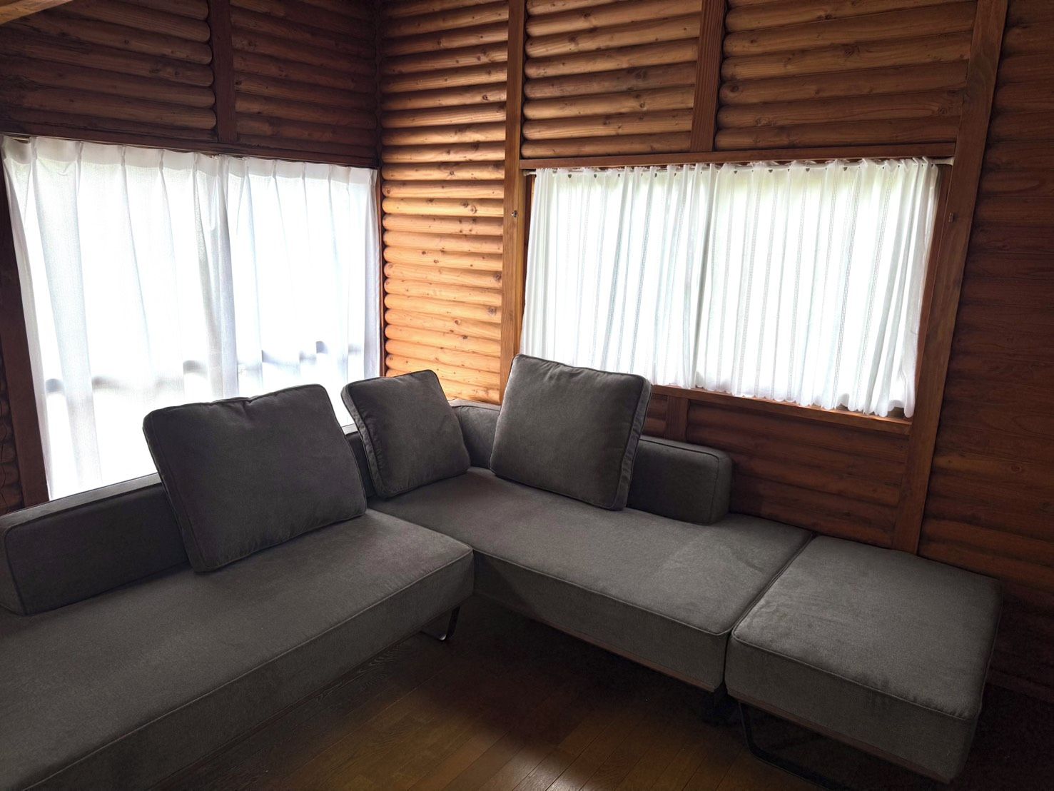 L’intérieur d’une cabane en rondins, avec un canapé d’angle, des murs en bois et de grandes fenêtres ornées de rideaux blancs.