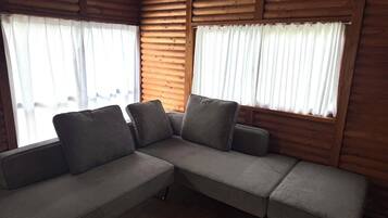 L’intérieur d’une cabane en rondins, avec un canapé d’angle, des murs en bois et de grandes fenêtres ornées de rideaux blancs.
