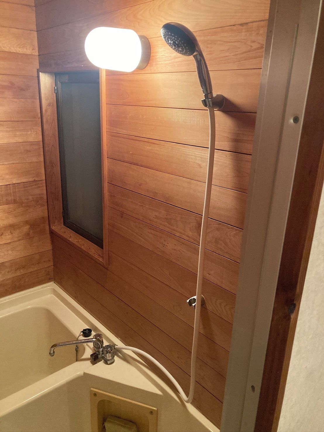 Une salle de bain aux murs lambrissés, équipée d’une baignoire, d’une pomme de douche et d’un luminaire fixé au mur.