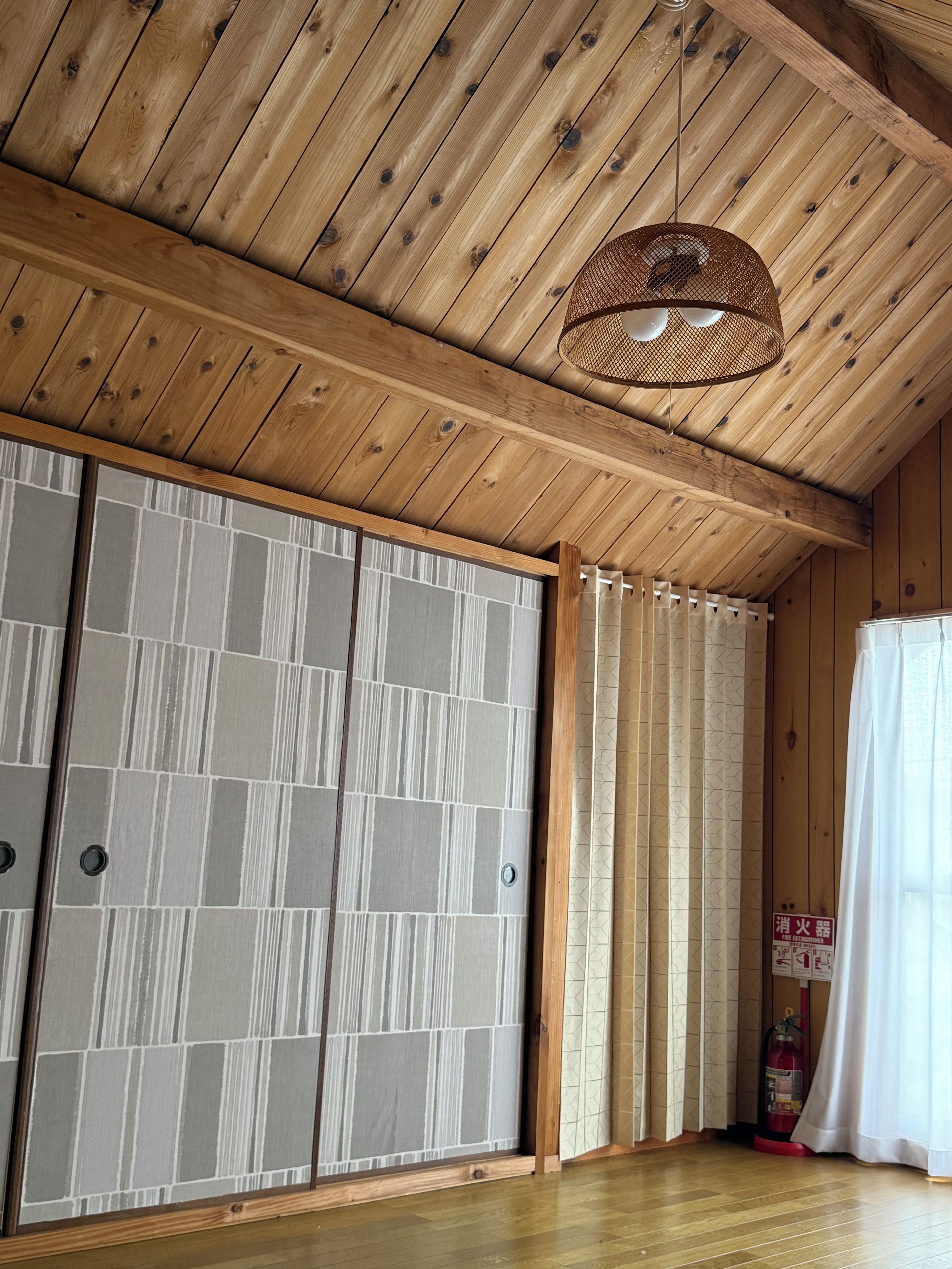 A room with wooden ceiling beams, a woven ceiling light fixture, sliding shoji screens, a fire extinguisher, and a curtain.