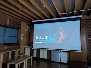 A room with a wooden ceiling, a large screen displaying a Netflix interface, a wooden bench, and a small refrigerator.