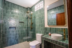 Un baño con azulejos verdes, inodoro blanco, ducha con cabezal dorado y lavamanos con grifo dorado.