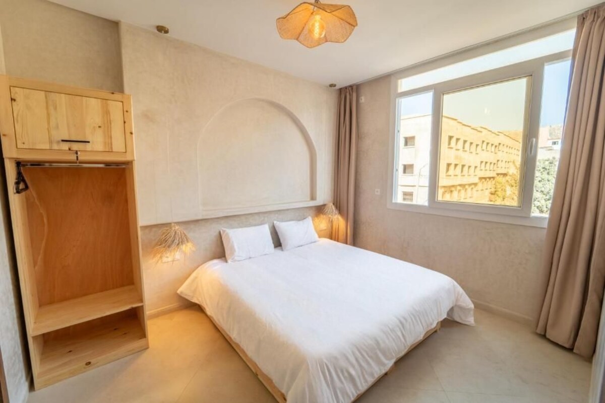 A bedroom with a bed, a wooden wardrobe, a window with a view, and a ceiling light fixture.