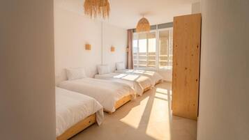 A bedroom with three beds aligned against the wall, a large window with curtains, and a wooden door.