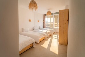 A bedroom with three beds aligned against the wall, a large window with curtains, and a wooden door. - Nomaya Surf House Tamraght (Aourir)