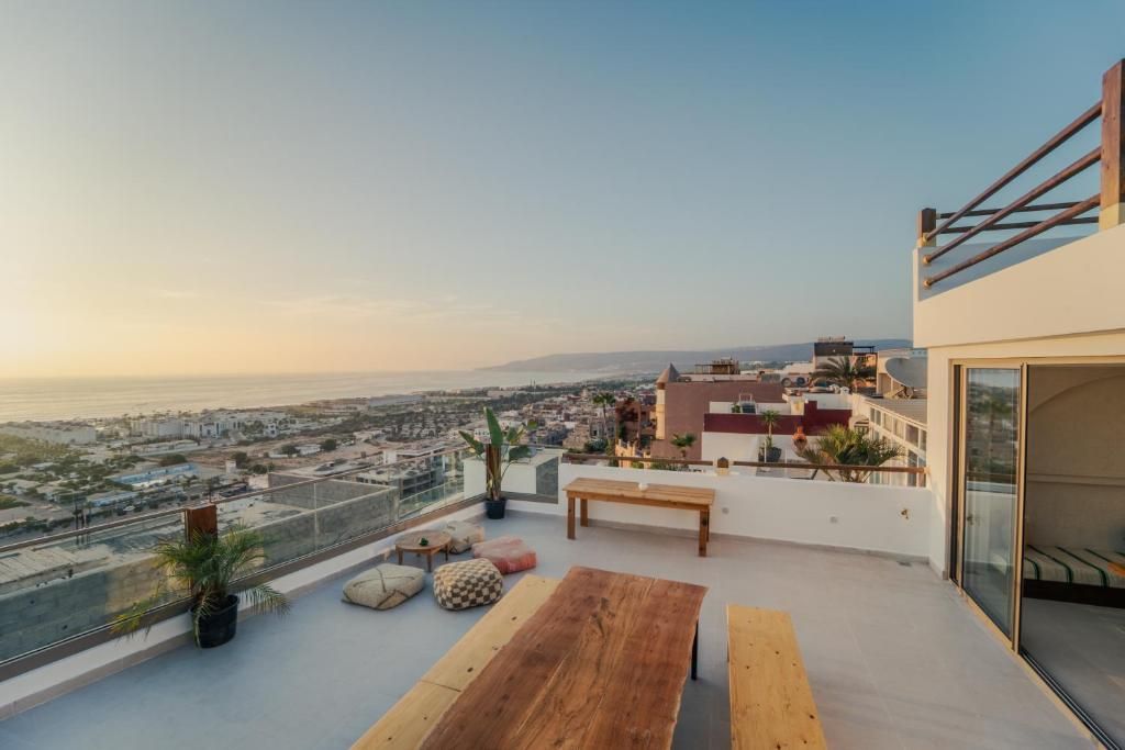 A rooftop terrace with a wooden table and benches, overlooking a cityscape and the sea.