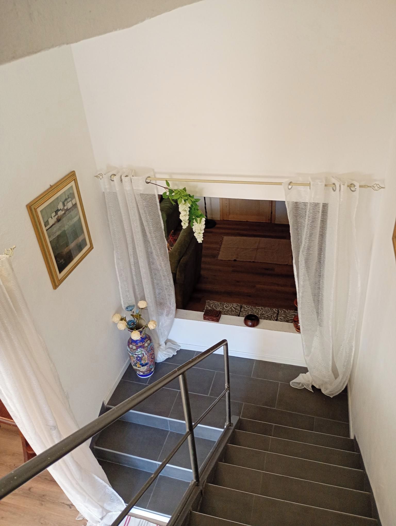 Una escalera con barandilla, cortinas blancas, un cuadro enmarco y un jarrón con flores.