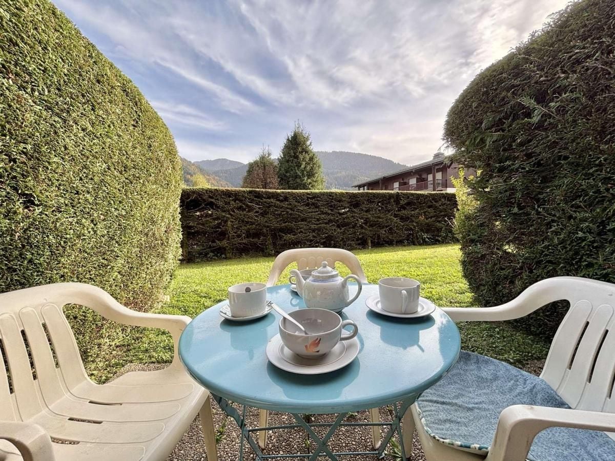 A round table with a blue tablecloth, surrounded by white plastic chairs, set outdoors with a view of neatly trimmed hedges and distant mountains.