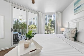 A hotel room with a bed, a bedside table with a tray, a lamp, and a view of palm trees and buildings. - Poolside Paradise Resort Pool (Destin)