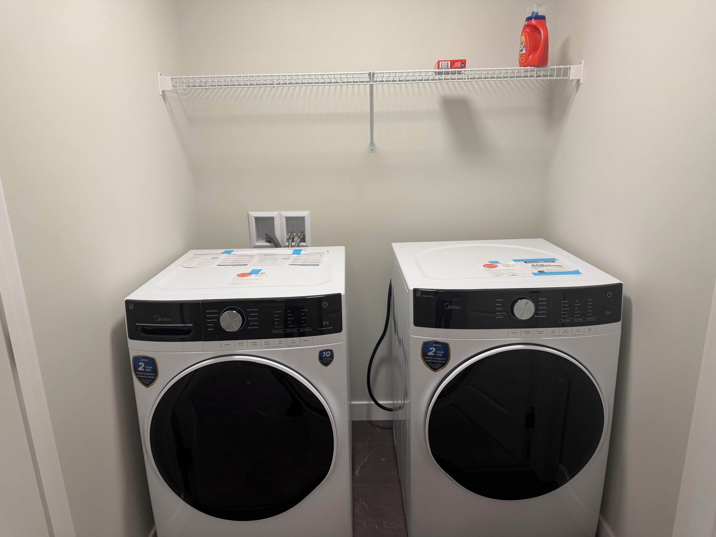 Two front-loading washing machines in a laundry room with a shelf above containing a bottle of detergent and a box.