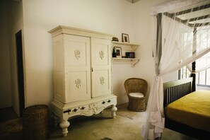 A bedroom with a bed, a white wardrobe, a wicker chair, and a shelf with books and a plant. - Oba Samaga by TONIK (Unawatuna)