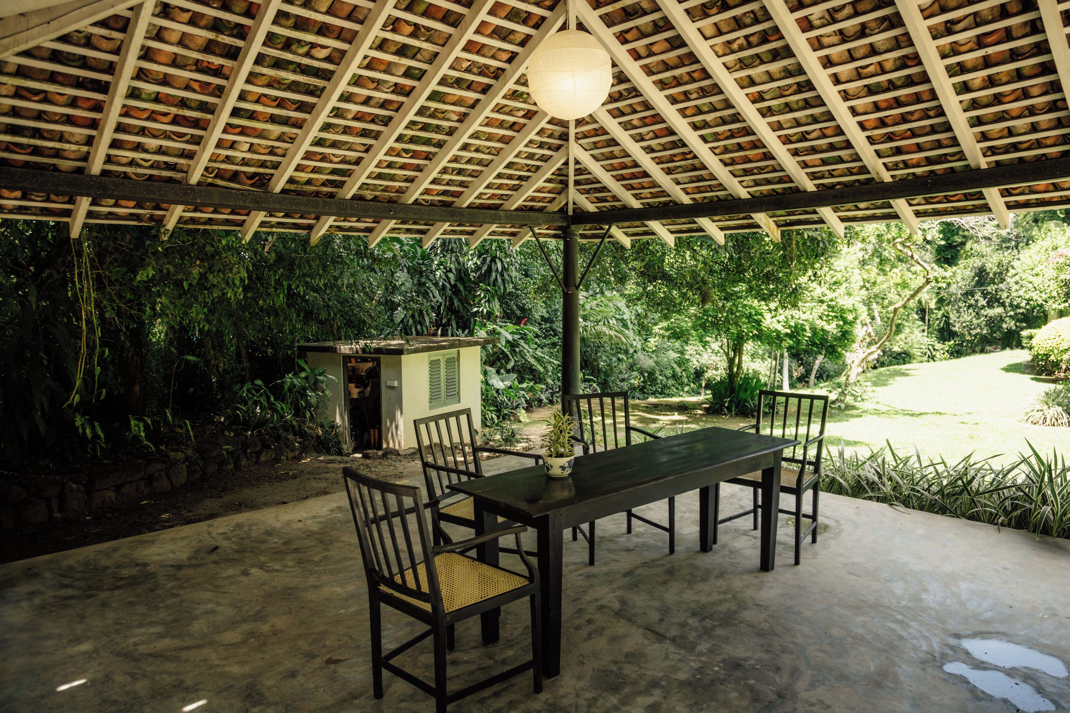 An outdoor covered patio with a table and chairs, surrounded by lush greenery.