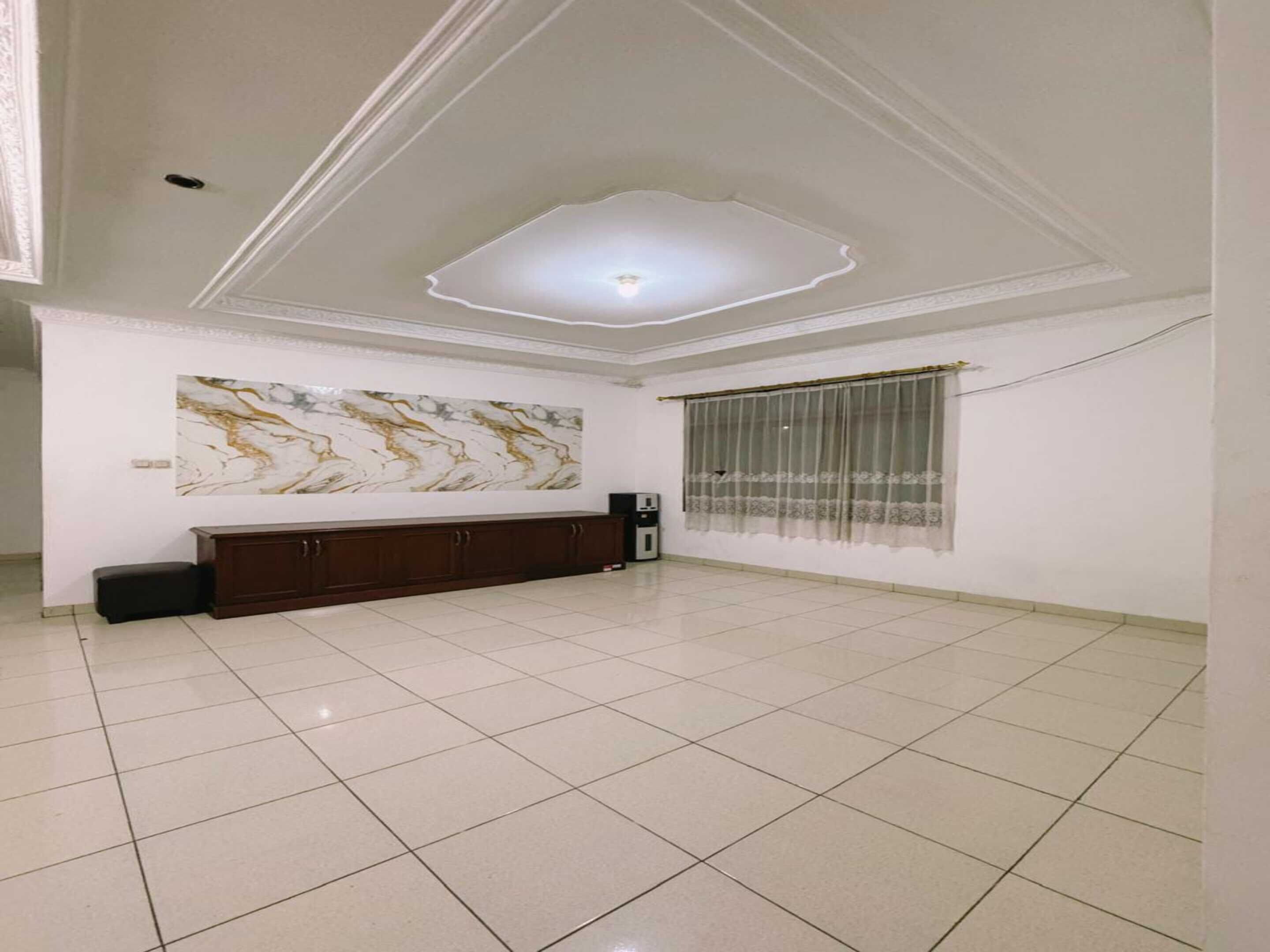 A spacious room with a tiled floor, a dark wooden cabinet, a marble wall art piece, and a window with sheer curtains.