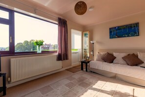 Une pièce bien éclairée, avec une grande fenêtre, un radiateur, un canapé, une table avec une lampe et un tableau accroché au mur.