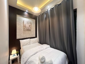 A neatly made bed with white linens and a rolled towel, a small bedside table with a lamp and a plant, and a large window with grey curtains.