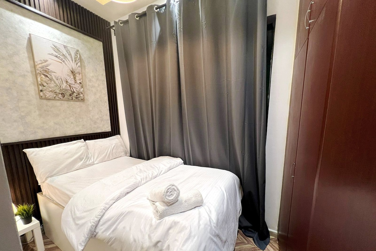 Een net opgemaakt bed met witte lakens en een opgerolde handdoek, een kleine potplant op een nachtkastje, en een schilderij aan de muur.