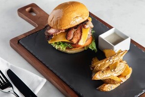 A burger with bacon, cheese, and lettuce on a bun, served with roasted potatoes and a small bowl of sauce on a black slate plate. - BonHomies Hotel (Port Harcourt)
