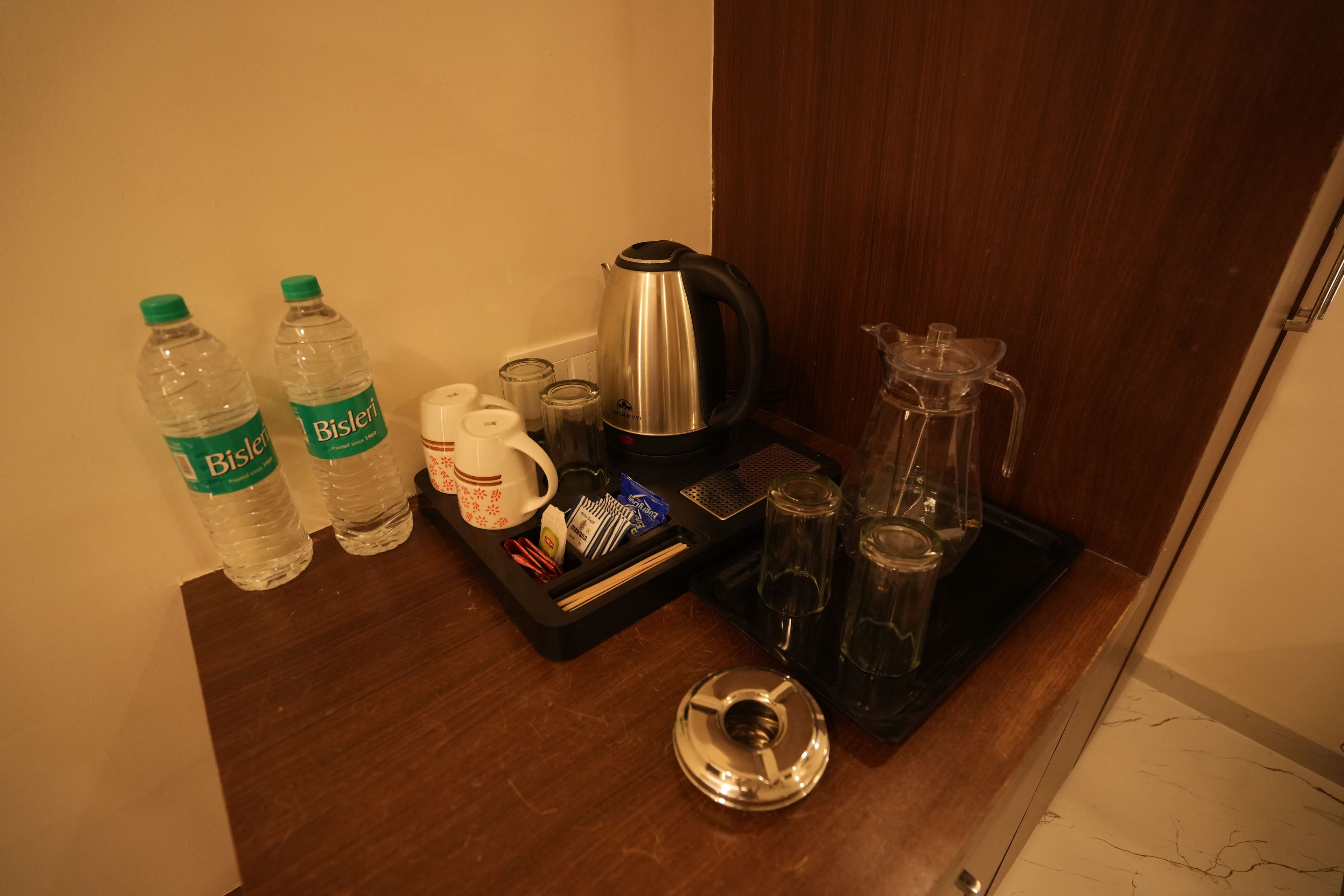 A kitchenette area with a coffee maker, two Bisleri water bottles, a mug, and a tray with utensils.