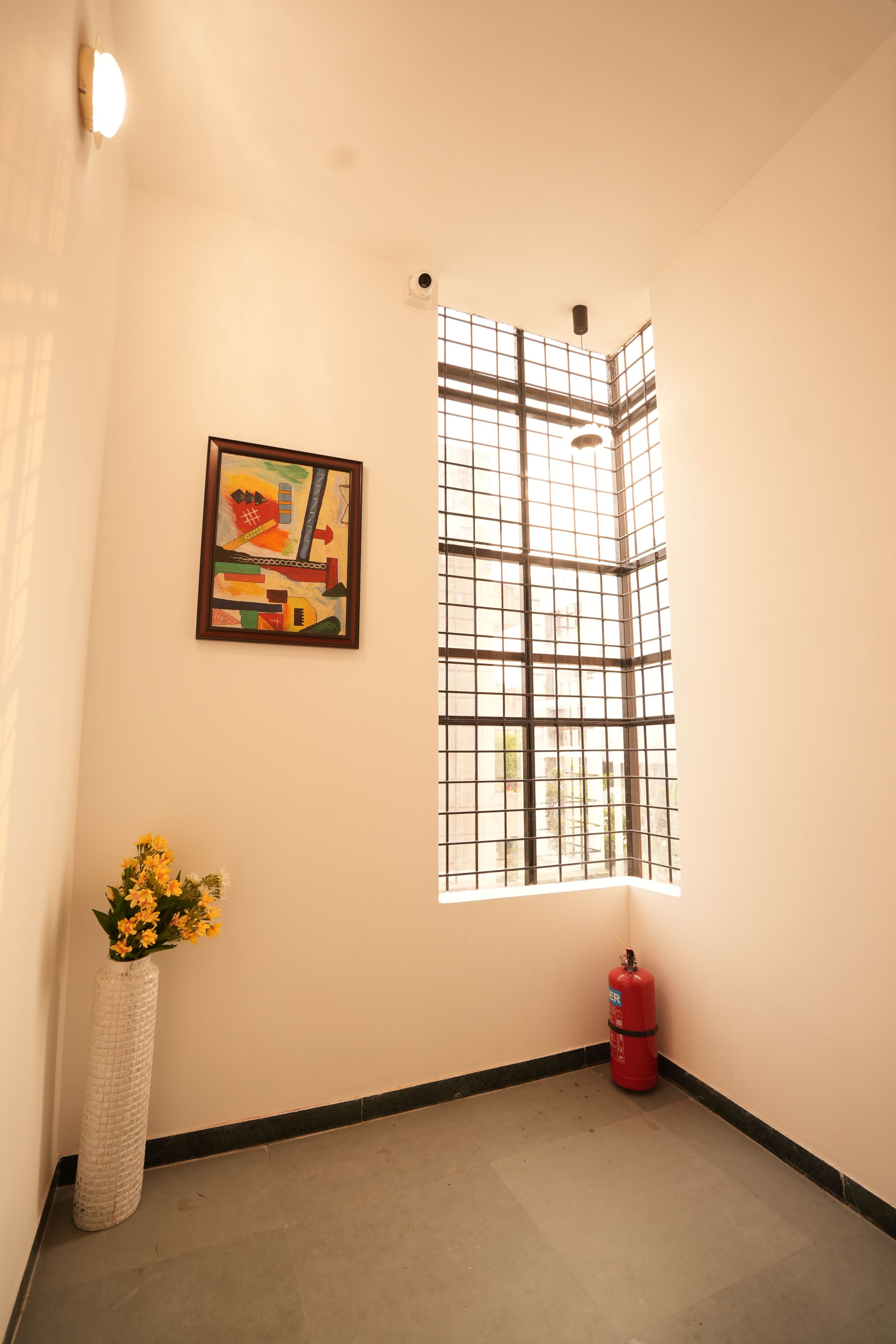Una habitación de esquina con ventana, un cuadro abstracto enmarco, un jarrón con flores y un extintor.