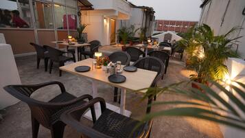 An outdoor dining area with tables and chairs, potted plants, and a building in the background.