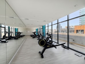 A gym with exercise equipment, large windows, and a view of the cityscape. - Bloomfields Studio in Azizi Riviera 43 (Dubai)
