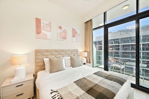 A modern hotel room with a large bed, a nightstand, a lamp, and a view of the city through large windows. - Bloomfields Studio in Azizi Riviera 43 (Dubai)