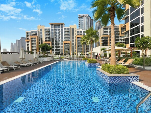 A swimming pool with tiled blue mosaic flooring, surrounded by lounge chairs and umbrellas, with high-rise buildings in the background. - Bloomfields Studio in Azizi Riviera 43 (Dubai)