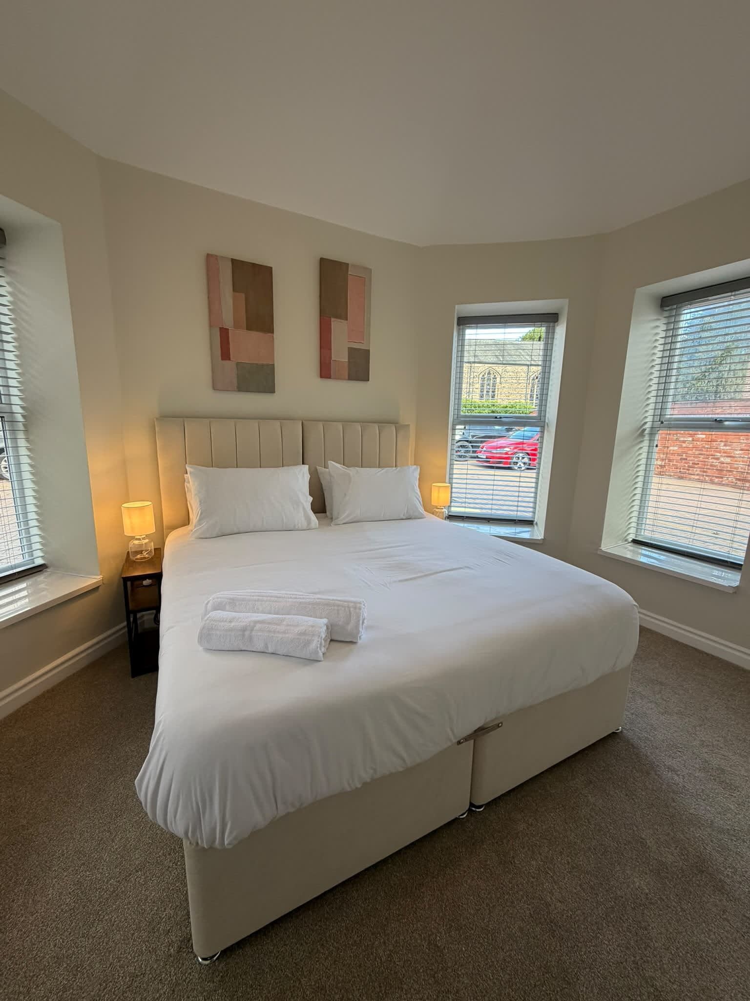 A bedroom with a large bed, two bedside lamps, and two windows with blinds.