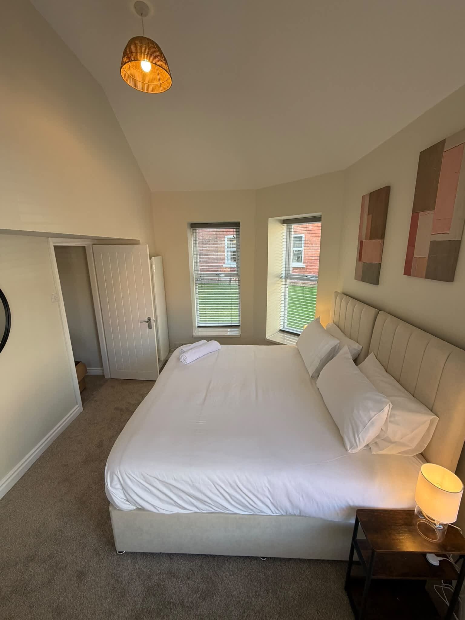 A bedroom with a large bed, two windows, a door, and a bedside table with a lamp.