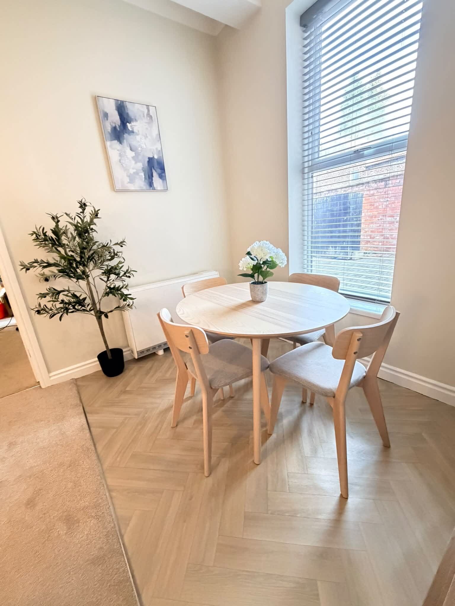 A dining area with a round table and four chairs, a potted plant, and a window with blinds.